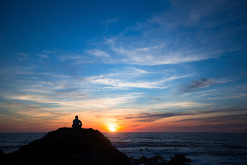 Silhouette of a lonely man sitting on the rocks at ocean beach during amazing sunset.