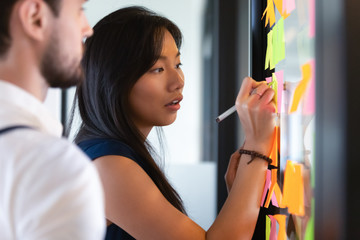 Focused asian businesswoman writing on sticky notes on glass