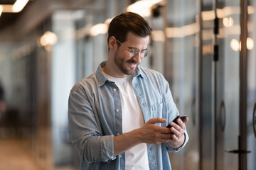 Happy young businessman using smart phone standing in office hallway