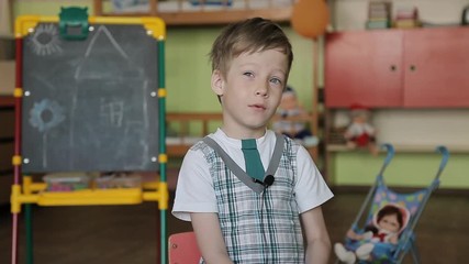 A 6 year old boy sits on a chair with a lapel microphone on his clothes and answers questions thoughtfully. The boy gives an interview in an educational envy against the background of a blackboard.