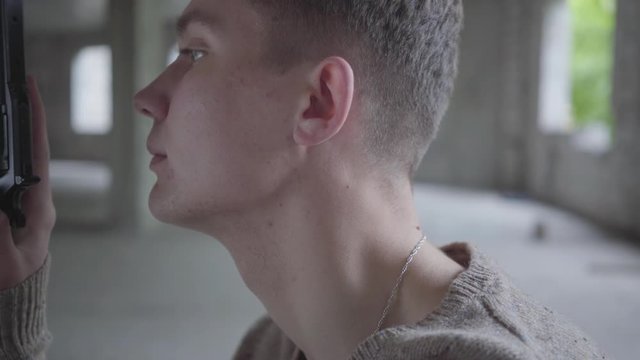Side view of a depressed crazy young man holding revolver in hands, slowly blowing out cigarette smoke in abandoned building. The guy going to commit suicide or kill human
