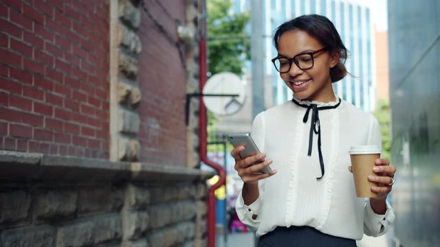 Smiling African American woman is using smartphone holding take out coffee walking outdoors in the street. People, devices and moden lifestyle concept.