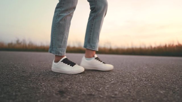 Unrecognizable woman in jeans and white sneakers walks along on asphalt road ahead. Concept of modern shoes, travel, hiking in summer nature.Camera shoots from right side.