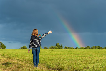 A woman with raised hands is standing in a field against the sky decorated with a rainbow after the rain.