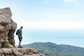 Young man with a backpack on top of cliff enjoying view of nature. Mountains and sea