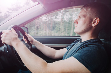 Young blond male driver is sitting behind the wheel. Close-up portrait. The man is wearing a dark blue plain t-shirt. Outside the window, the trees are blurry due to the movement of the car.