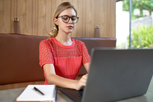 Closeup lifestyle portrait of professional blonde caucasian millennial student wearing glasses taking notes on laptop computer in a modern restaurant