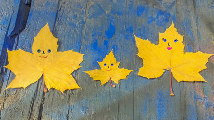 three yellow maple leaves with cheerful muzzles painted on them lie on a blue wooden surface