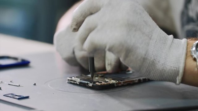 Repairing mobile phone in workshop, hands closeup. Repairman in white gloves disassemble smartphone with screwdriver in service center. Master in white gloves unscrews bolts. Repair and maintenance.