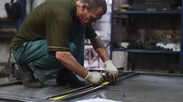Man worker locksmith measures tape measure length of metal bars. He makes marks with pencil in workshop. He is in uniform and gloves in his workplace. Work with metal in production.