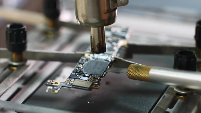 Repairman inserts knife under chip's part heating it with dryer. Removing chipset element microprocessor using knife melted with soldering dryer. Electronics engineer repairing chip in workshop.