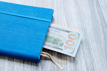 A hundred-dollar bill in a blue notebook with a blue ribbon. Background grey wooden surface. The concept of calculating the budget for the business, opening an account for payment.