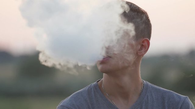 Handsome stylish young man with long brunette hair vaping outdoors at sunset in hot summer day at sunset, Close up 