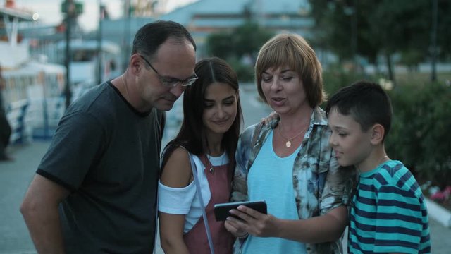 Positive family watching their cool photos on smartphone outdoors in the evening on the quay. Four people with a phone