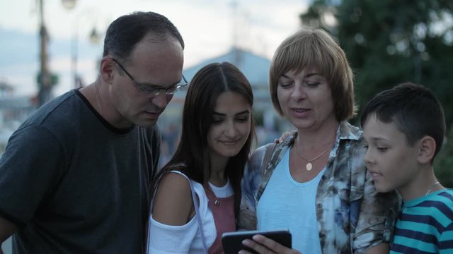 Positive family watching their cool photos on smartphone outdoors in the evening on the quay. Four people with a phone