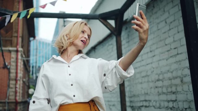 Slow motion of pretty middle aged woman taking selfie with smartphone outdoors posing for mobile camera having fun. People, devices and photo concept.