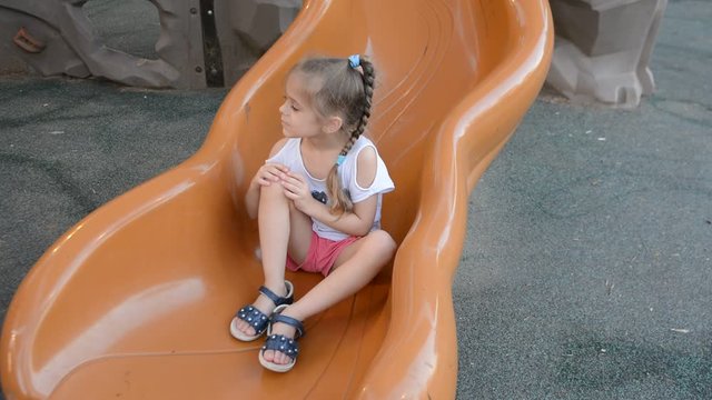 Little girl sitting on a top of a slide and smiling. Baby on the playground in the park.