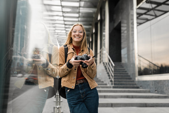 Portrait of professional photographer in stylish eyeglasses holding digital camera outdoors. Smiling hipster woman with beautiful emotional face walking around urban street. Successful hobby 