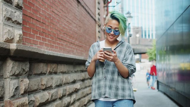 Slow motion of Asian hipster walking using smartphone and holding to go coffee outdoors in city street. Modern technology, youth and urban lifestyle concept.
