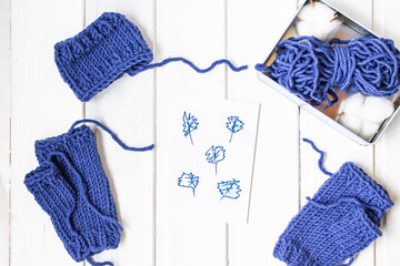 Knitted mittens withiout fingers. Woolen yarn ball. White wooden background. Copy space. Place for text. Top view.  Metal box with cotton. Beautiful card with blue leaves