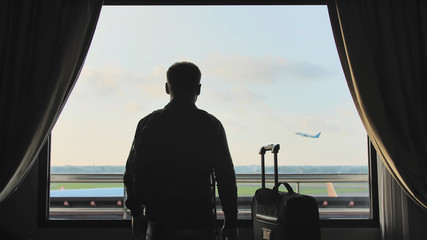 A young guy looks at the plane taking off from the window of his hotel room and leaves with a suitcase for boarding.