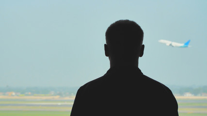Silhouette of a young man watching the take-off of an airplane against a blue sky.