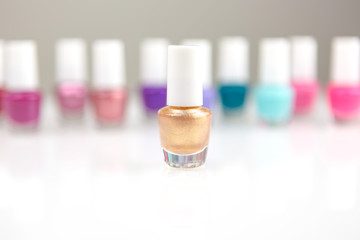 View on a shiny white desk full of small glass and plastic bottles with assorted nail polish and lacquer on a back creating row with focus on one golden nail polish.