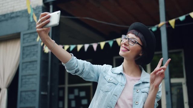 Portrait of attractive young brunette taking selfie outdoos with smartphone posing for camera standing outside in city street. People and photo concept.