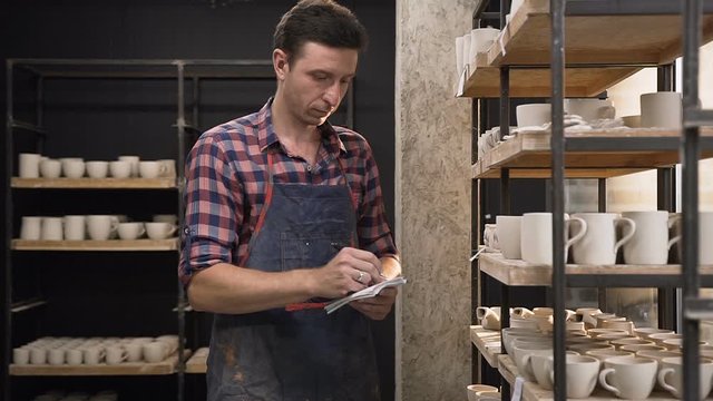 Focused male potter counting cermics from the shelves and noting in the notebook.