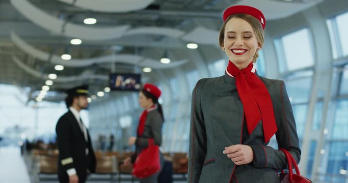 Portrait shot of the young blonde pretty Caucasian stewardess in the stylish uniform standing in the airport hall and smiling happily.