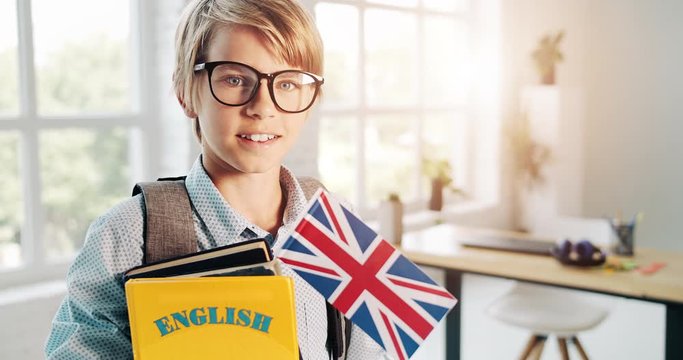 Cute schoolboy in eyeglasses holding english book and british flag, looking to camera and smiling, education