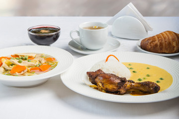 Delicious lunch with chicken leg, rice, peas, soup, dessert, tea and bread. Served on a white table.