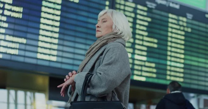 Rear of the caucasian senior woman with gray hair standing in the airport terminal and looking at the flight timetable, then turning to the camera. Back view.