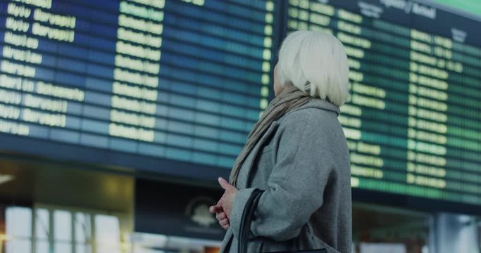 Caucasian old lady with gray hair standing in the airport terminal and then turning to look at the flight timetable. Back view. Rear.