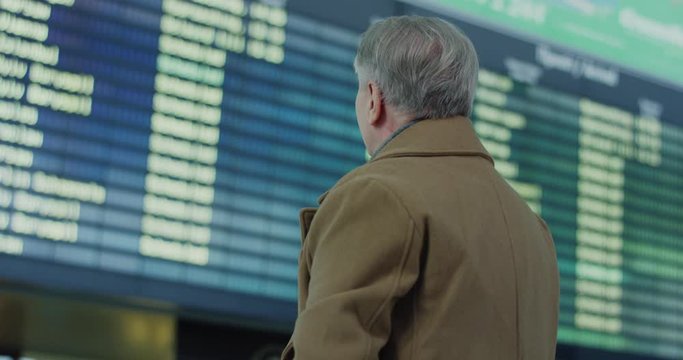 Caucasian old man looking at the watch on his hand while standing in the airport on the flight timetable background.