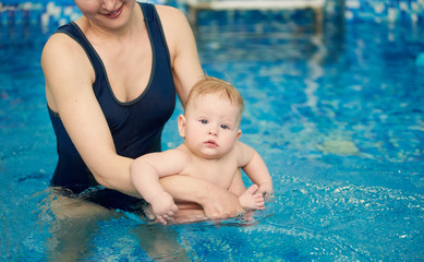 Cropped snapshot of young smiling mother holding her son sitting in her arms in pool in blue water. Baby looking in camera. Infant swimming and doing water exercises. Health promotion concept