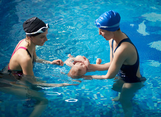 Side view of mother and female young instructor carrying out swimming lessons for parent and baby. Mom and her newborn child at infant swimming class. Baby floating lying on his back in blue water