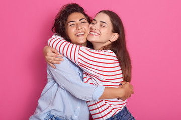 Close up portrait off positive ladies, frienads or sisters standing next to each other, having warm hug, posing with pleasant smiles, isolated over pink background. Friendship and happyness concept.