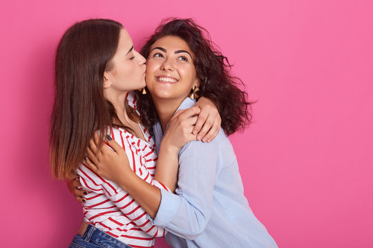 Image of two cheerful young girls in vivid clothes hugging and kissing on cheek, isolated on rosy wall background, women in love spend time together. People sincere emotions and same sex love concept.