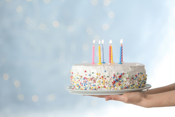 Woman holding birthday cake with burning candles against blurred background, closeup. Space for text