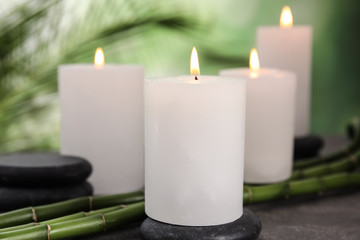 Fototapeta na wymiar Burning candles, spa stones and bamboo sprouts on grey table against blurred green background