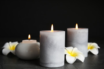 Fototapeta na wymiar Burning candles and plumeria flowers on dark grey table against black background