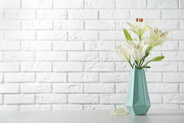 Vase of beautiful lilies on table against  white brick wall, space for text