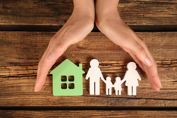 Woman holding hands near figures of house and family on wooden background, top view