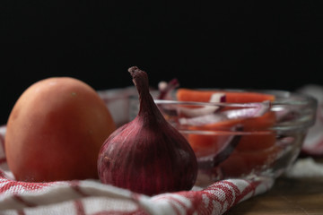 Close-up of tomato and pink onion, and a salad of them in a glass bowl with the addition of vegetable oil, handful of sea salt and a red on white napkin on a wooden table 