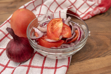 A salad of fresh tomatoes and pink onion in a glass bowl with the addition of vegetable oil, handful of sea salt and a red on white napkin on a wooden table on a black background. Side view