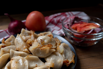 Hot, boiled and fried with onions, lard, dumplings with potatoes and a salad of tomatoes and pink onion with vegetable oil and a red and white napkin on a wooden table on a black background