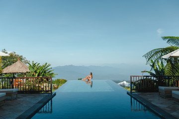 Slim young woman in swimsuit relaxing on edge tropical infinity pool in mountains. Palms around and crystal clean water. Luxury resort on Bali island