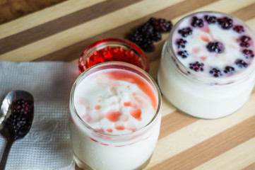 Yogurt in transparent jars with fresh blackberry and strawberry voyen with a spoon, on wooden background