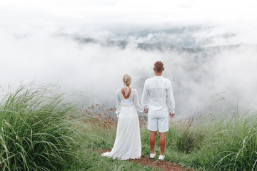 Fototapeta premium Newlyweds walk and hold hands during honeymoon trip in mountains. Back view.The groom embraces the bride from the back side on the rocks with a magnificent view